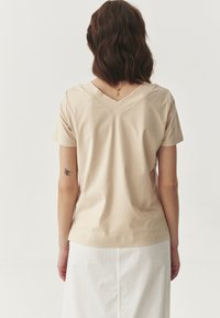 Beige, rövid ujjú, V nyakú felső, puha anyagból készült, sima felülettel. Enyhe fényességgel és laza szabással rendelkezik, egy világos szoknyába tűrve.