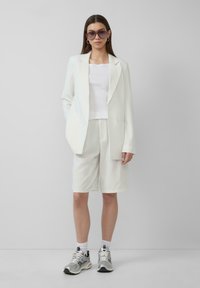 Witte blazer en short set met streepjes, gecombineerd met een witte top en zilveren sneakers. Overdreven zonnebril maakt de look compleet.