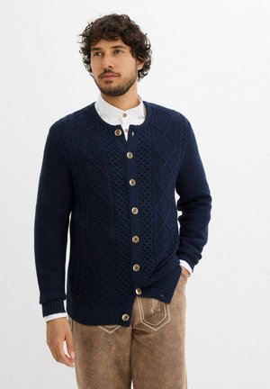 Marineblauer gestrickter Cardigan mit einem Rautenmuster, Rundhalsausschnitt und großen Holzknöpfen, kombiniert mit hellen Hosen.