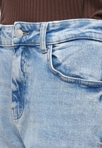 Jeans de ganga azul claro com acabamento desbotado, apresentando fecho com botão de metal e design clássico de cinco bolsos. Tecido texturado com costuras visíveis.