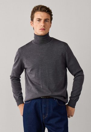 Hombre de pie frente a un fondo liso, con un jersey gris de cuello alto y vaqueros azules, mirando al frente con expresión neutral.