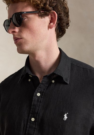 Νέος άνδρας με σγουρά μαλλιά που κοιτάζει αριστερά, φοράει στρογγυλά μαύρα γυαλιά ηλίου και μαύρο πουκάμισο με κουμπιά και λευκό λογότυπο παίκτη πόλο.