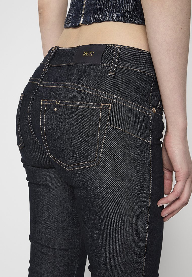 Jeans blu scuro con un design aderente, caratterizzati da cuciture arancioni e una piccola etichetta con logo sulla vita. Stile a cinque tasche, classica trama in denim.