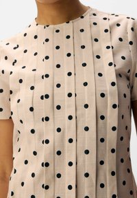 Beige kortærmet bluse med sorte polkaprikker og lodrette plisseeffekter på forsiden, båret af en person synlig fra halsen til taljen.