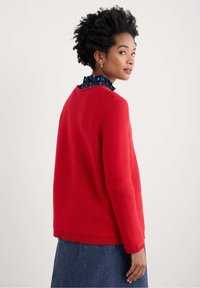 Pull en tricot rouge à côtes avec un col rond et des manches longues, superposé à une blouse bleu marine à motifs, associé à une jupe en denim.