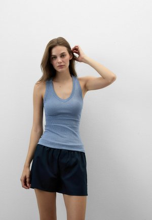 Jonge vrouw met lang bruin haar, gekleed in een blauwe mouwloze top en zwarte korte broek, staand tegen een effen witte muur.