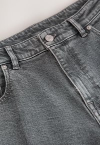 Pelēku džinsu vidukļa daļas tuvplāns, kur redzams pogas, jostas cilpas, priekšējās kabatas un šūšanas detaļas.