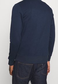 Námořnická modrá mikina s kulatým výstřihem, žebrovými manžetami a lemem; vyrobena z texturované látky, nošena s tmavými džínami.