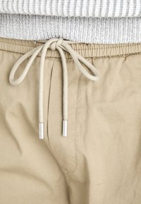 Beige drawstring pants with a soft texture, featuring a grey ribbed waistband and metal-tipped drawstrings for adjustable fit.