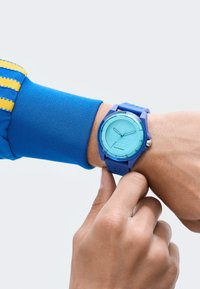 Sinine silikoonkell ümmarguse helesinise kellaga. Omadusteks on tekstuuriga rihm ja minimaalsed tunnimärgid, küljel on metallist kroon.