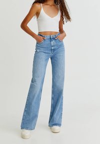 Jeans de tiro alto en azul claro con pierna acampanada, que presentan una textura desvaída y desgastes en las rodillas, combinados con una camiseta corta blanca.