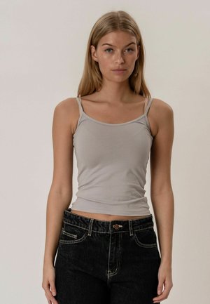 Jeune femme aux cheveux blonds raides portant un débardeur gris clair à fines bretelles et un jean en denim noir, debout devant un fond uni.