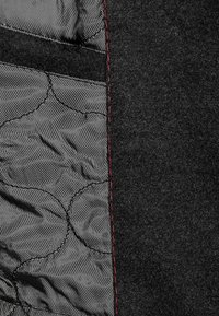 Schwarzer Wollstoff mit einem gepolsterten Futter, das ein strukturiertes Muster aufweist. Rote Nähte entlang des Randes setzen einen kontrastierenden Akzent.