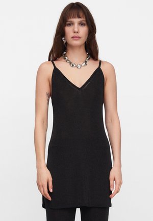 Frau trägt ein schwarzes, ärmelloses V-Ausschnitt-Kleid mit dünnen Trägern und eine auffällige silberne Halskette, steht vor einem schlichten weißen Hintergrund.