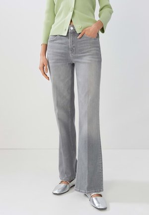 Person iført lys grøn ribbet cardigan og højttaljede grå flare jeans med sølv firkantede sko, hånd i lommen.