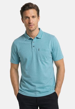 Mężczyzna ubrany w zielononiebieską koszulkę polo z krótkim rękawem i kieszenią na piersi, stojący z rękami w kieszeniach czarnych spodni na jednolitym tle.