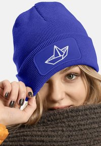 Blue knitted beanie with a textured finish and a patch featuring a white paper boat design. Soft fabric, stretchable fit.