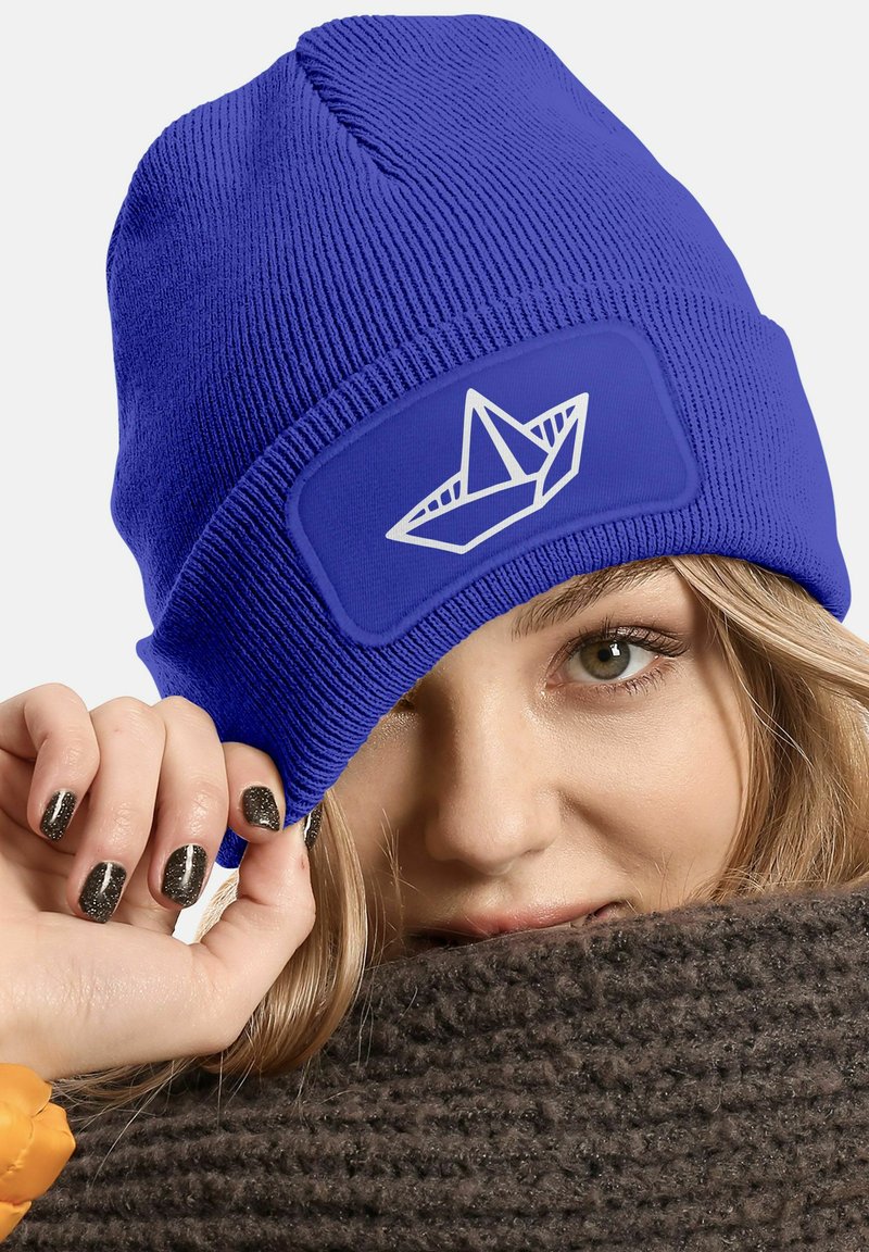 Blue knitted beanie with a textured finish and a patch featuring a white paper boat design. Soft fabric, stretchable fit.