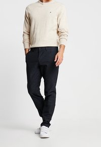 Beżowy sweter z długim rękawem i okrągłym dekoltem z prążkowanymi mankietami, zestawiony z ciemnoszarymi, dopasowanymi spodniami i białymi trampkami, prezentujący casualowy wygląd.