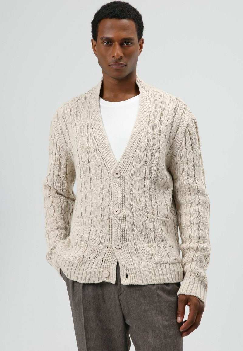 Beige gebreid vest met kabelpatroon, V-hals, lange mouwen en voorzakken, afgewerkt met knopen en een zachte textuur.