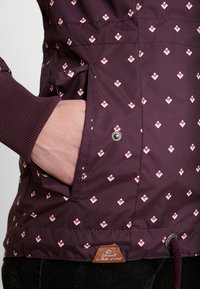 Hand i fickan på en vinröd jacka med små vita och röda geometriska mönster och ribbad manschett, bärd över svarta byxor.