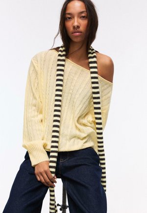 Donna che indossa un maglione giallo pallido a trecce con le spalle scoperte, jeans scuri e una lunga sciarpa a righe seduta su uno sgabello contro uno sfondo bianco.