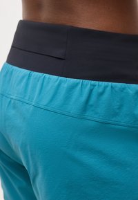 Κοντινό πλάνο ενός ατόμου που φοράει μπλε παντελόνι με φαρδιά μαύρη ελαστική μέση, μπροστά σε απλό φόντο.