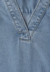 Lyseblå denimstof med en V-formet halsudskæring med dobbeltsyede sømme og subtile teksturvariationer.