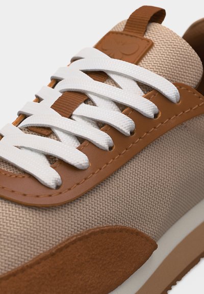 Μπεζ και καφέ αθλητικό παπούτσι με λευκά κορδόνια, υφασμάτινο επάνω μέρος με υφή, δερμάτινα περιγράμματα και θηλιά για τράβηγμα στη γλώσσα.