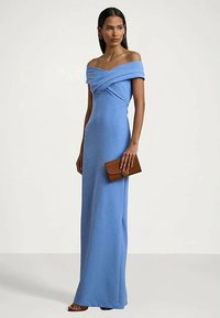 Γυναίκα που φοράει έναν μακρύ, off-shoulder μπλε φόρεμα κρατώντας μια καφέ τσάντα, στέκεται μπροστά σε μια απλή ανοιχτή φόντο.