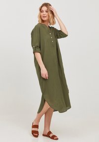 Robe verte olive à manches longues avec un décolleté en V, patte de boutonnage, manches retroussées et fentes sur les côtés. Portée avec des sandales marron.