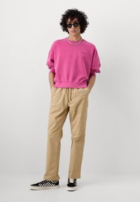 Lyserød trøje med ribbet manchetter, beige løstsiddende bukser og sorte ternede sneakers. Modellen bærer solbriller og en kæde-halskæde.
