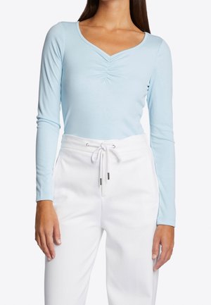 Kobieta ubrana w jasnoniebieski, długi rękaw z marszczonym dekoltem w kształcie litery V oraz białe spodnie ze sznurkiem, na tle jednolitego tła.