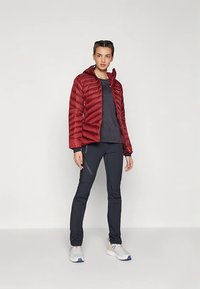 Raudona pūkinė striukė su siuvinėta tekstūra ir užtrauktuku, derinama su tamsiomis sportinėmis kelnėmis ir baltais sportbačiais, neutralaus fono kontekste.