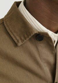 Cuello de chaqueta de pana marrón con un botón negro, superpuesto a una camisa blanca con rayas beige. Tejido texturizado con costuras visibles.