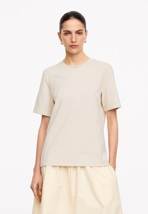 Frau mit dunklen Haaren in einem glatten Dutt trägt Perlenohrringe, ein beigefarbenes Kurzarm-T-Shirt und cremefarbene weite Hosen vor weißem Hintergrund.