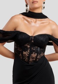 Musta iltapuku, jossa on pitsinen bodice, sydänmainen kaula-aukko ja tiukka hame. Läpinäkyvä musta huivi hartioiden ympäri. Suuret koristeelliset korvakorut.