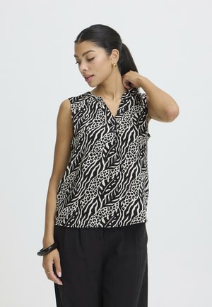 Donna con top nero e bianco senza maniche con stampa animalier, pantaloni neri, orecchini dorati, bracciale nero, che posa con la mano vicino al collo.