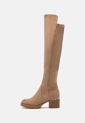 Botte haute en daim beige avec talon bloc, semelle crantée et léger accent doré au-dessus du talon, vue de côté.