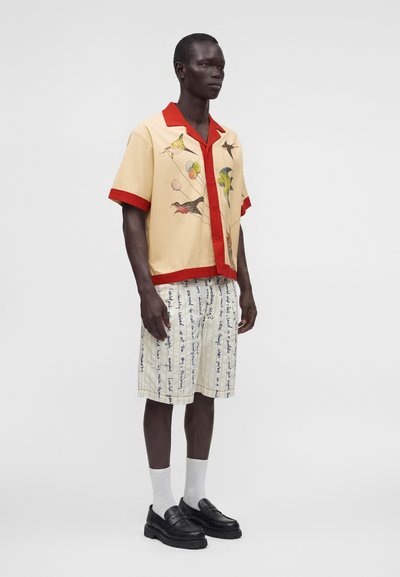 Homme grand portant une chemise beige à manches courtes avec une bordure rouge et des imprimés d’oiseaux, un short blanc avec un texte bleu, des chaussettes blanches et des mocassins noirs.