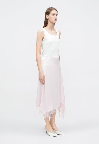 Lehké bílé tílko bez rukávů v kombinaci s asymetrickou světle růžovou sukní s krajkovým lemem; modelka má na nohou bílé pantofle na podpatku.