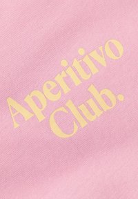 Κίτρινο κείμενο «Aperitivo Club.» τυπωμένο σε απαλό ροζ υφασμάτινο φόντο, δείχνοντας την υφή του υφάσματος και λεπτομέρειες στενής λήψης.