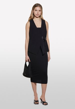 Gilet sans manches noir avec décolleté en V et ceinture à la taille, associé à une jupe midi noire ajustée. Le mannequin tient un sac à main noir texturé et porte des sandales à talons noires.