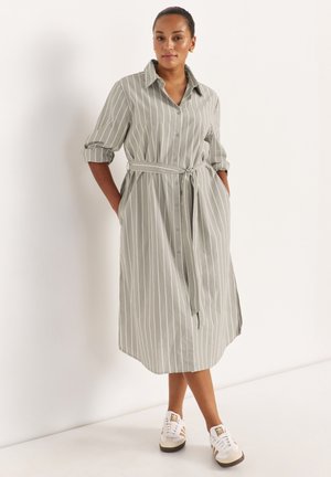 Frau trägt ein langärmeliges Hemdblusenkleid mit grauen und weißen vertikalen Streifen, Gürtel, hochgekrempelte Ärmel und weiße Turnschuhe, steht vor einer schlichten Wand.