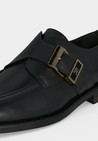 Chaussure de ville en cuir noir avec une boucle métallique dorée présentant un logo sur le côté, des coutures visibles et un talon bas empilé.
