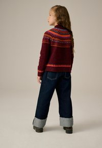 Sweter w kolorze buraczkowym wykonany na drutach, ozdobiony wielokolorowymi wzorami geometrycznymi na karczku, zestawiony z ciemnoniebieskimi jeansami z podwiniętymi nogawkami i czarnymi butami.