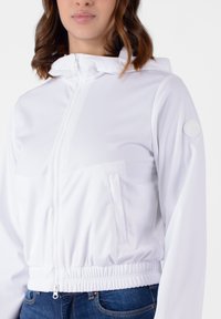 Giacca bianca con zip e cappuccio, dotata di orlo elastico, presenta una tasca frontale e una patch in tessuto con logo sulla manica sinistra.
