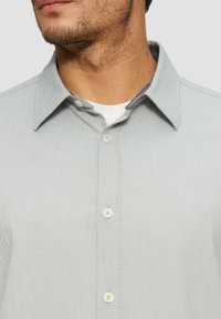 Helles grau button-up Hemd mit einem strukturierten Muster, ausgestattet mit einem klassischen Kragen und weißen Knöpfen, über einem weißen Unterhemd getragen.