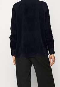 Top in velluto a coste blu navy con maniche lunghe e polsini a bottoni, caratterizzato da una vestibilità rilassata e una texture a coste. Abbinato a pantaloni neri a gamba larga.