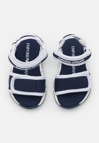 Námornícke modré sandále s bielymi akcentmi; majú nastaviteľné popruhy a logo. Vyrobené z odolného materiálu s textúrovanou podrážkou.